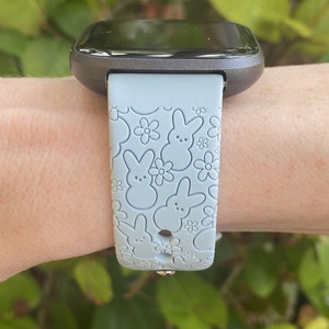 K&ouml;nnte beinhalten: Eine graue Smartwatch mit einem hellblauen Armband mit einem Muster aus Hasen- und Blumenmotiven. Die Uhr wird am Handgelenk getragen, mit gr&uuml;nem Laub im Hintergrund.