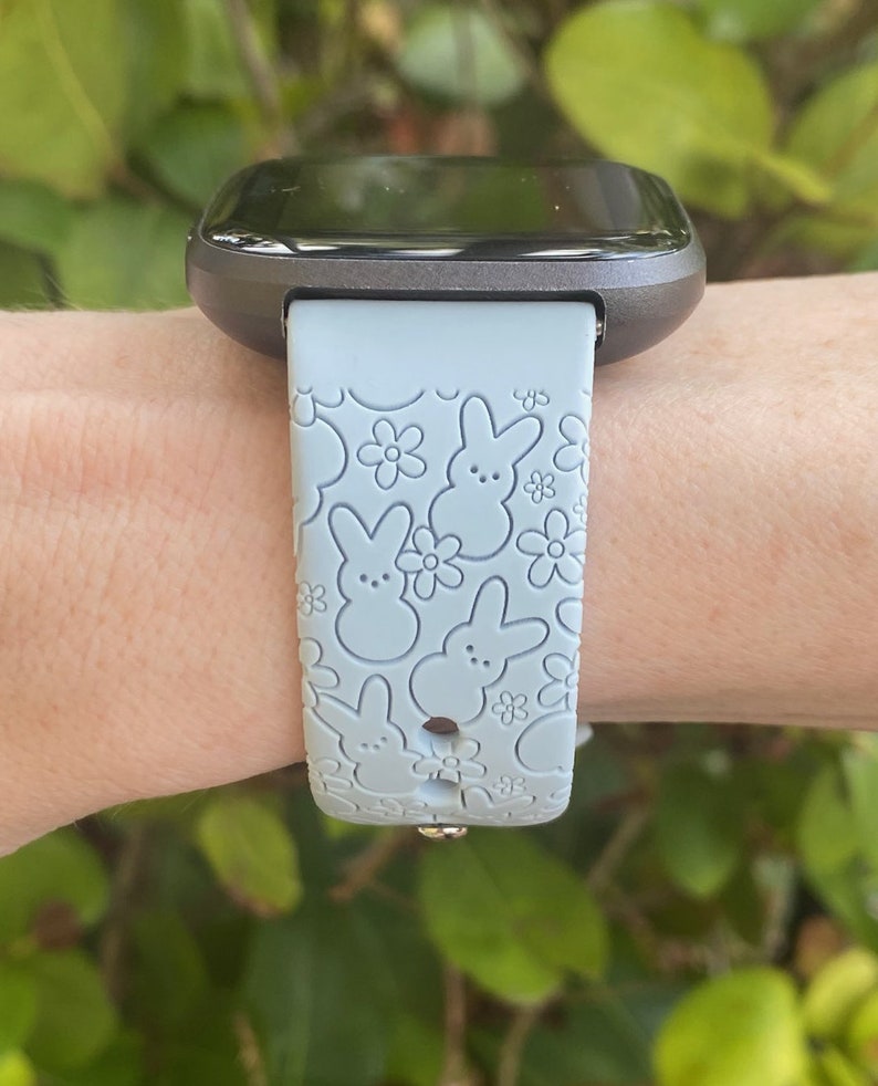 K&ouml;nnte beinhalten: Eine graue Smartwatch mit einem hellblauen Armband mit einem Muster aus Hasen- und Blumenmotiven. Die Uhr wird am Handgelenk getragen, mit gr&uuml;nem Laub im Hintergrund.