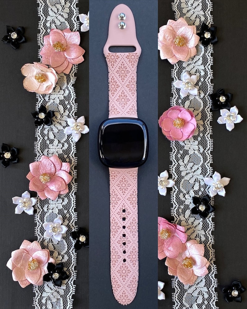 Puede incluir: Una banda de reloj inteligente rosa con un delicado patr&oacute;n floral. La banda tiene una hebilla plateada y se muestra sobre un fondo negro con ribete de encaje.