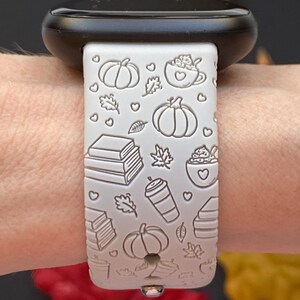 May include: A light gray Apple Watch band with a fall theme. The band features embossed pumpkins, leaves, books, and coffee cups. The watch case is black.