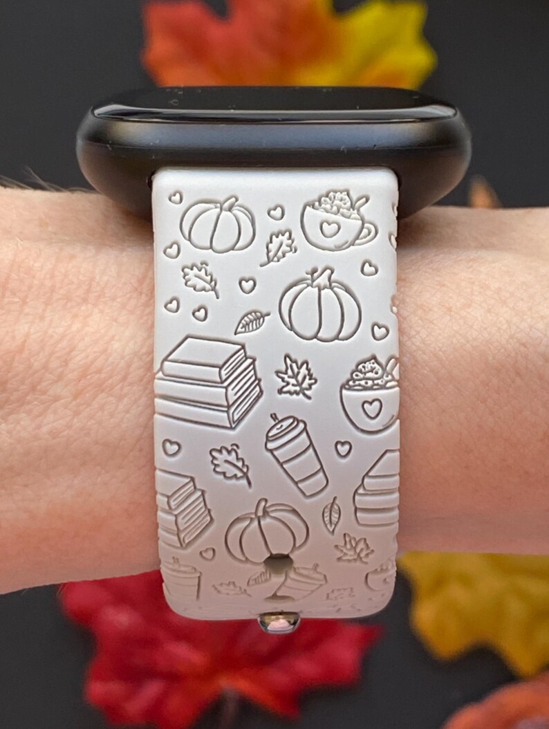 May include: A light gray Apple Watch band with a fall theme. The band features embossed pumpkins, leaves, books, and coffee cups. The watch case is black.