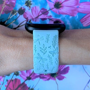 May include: A light blue smartwatch band with a floral pattern featuring dragonflies and leaves. The band is made of silicone and has a black metal buckle.