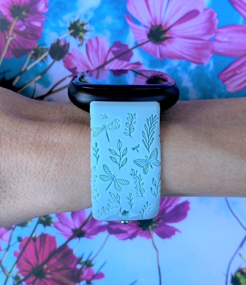 May include: A light blue smartwatch band with a floral pattern featuring dragonflies and leaves. The band is made of silicone and has a black metal buckle.