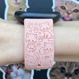 K&ouml;nnte beinhalten: Rosa Smartwatch-Armband mit gepr&auml;gten Cartoon-Katzen und Pfotenabdr&uuml;cken. Das Armband ist an einer schwarzen Smartwatch befestigt. Das Design umfasst verschiedene Katzenposen und ein Fischskelett. Ein lustiges Accessoire f&uuml;r Katzenliebhaber.