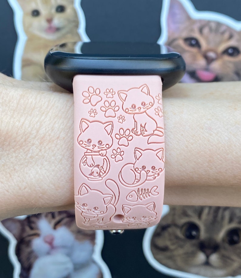 K&ouml;nnte beinhalten: Rosa Smartwatch-Armband mit gepr&auml;gten Cartoon-Katzen und Pfotenabdr&uuml;cken. Das Armband ist an einer schwarzen Smartwatch befestigt. Das Design umfasst verschiedene Katzenposen und ein Fischskelett. Ein lustiges Accessoire f&uuml;r Katzenliebhaber.
