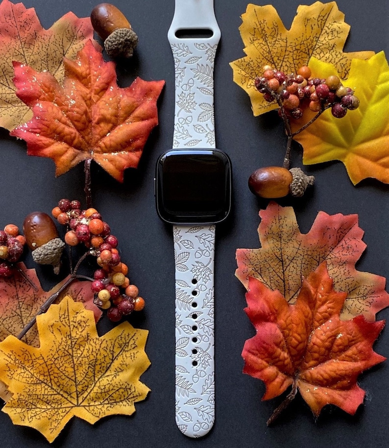 May include: A smartwatch with a light gray band featuring a fall leaves and acorn pattern. The watch is surrounded by colorful fall leaves in shades of red, orange, and yellow, along with acorns and berries, all set against a black background.