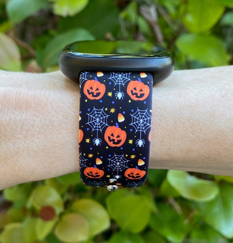 May include: A black smartwatch with a Halloween-themed band. The band features a repeating pattern of orange pumpkins, spiderwebs, and candy corn on a black background.