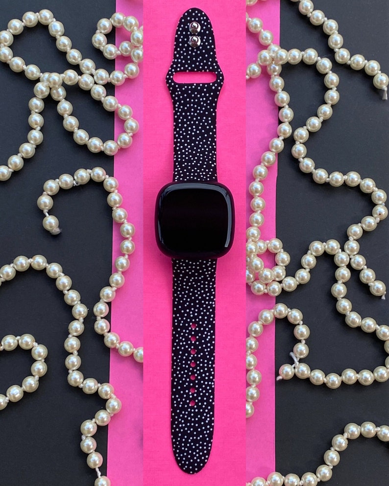 Pu&ograve; includere: Un orologio intelligente nero con un cinturino a pois bianchi su uno sfondo rosa. L'orologio &egrave; circondato da collane di perle.