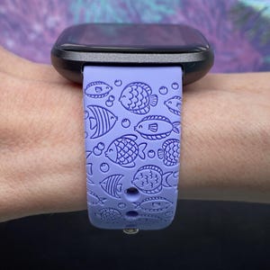 May include: A purple silicone watch strap with a fish pattern. The strap is designed for a smartwatch and has a black watch face.