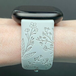 May include: White smartwatch band with a floral design. The band is made of silicone and has a raised floral pattern.