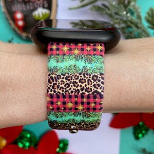 Op de afbeelding: Een zwarte smartwatch met een kleurrijk kerstbandje. Het bandje heeft een rood-zwart ruitpatroon, een groen-gouden bladpatroon en een luipaardprint.