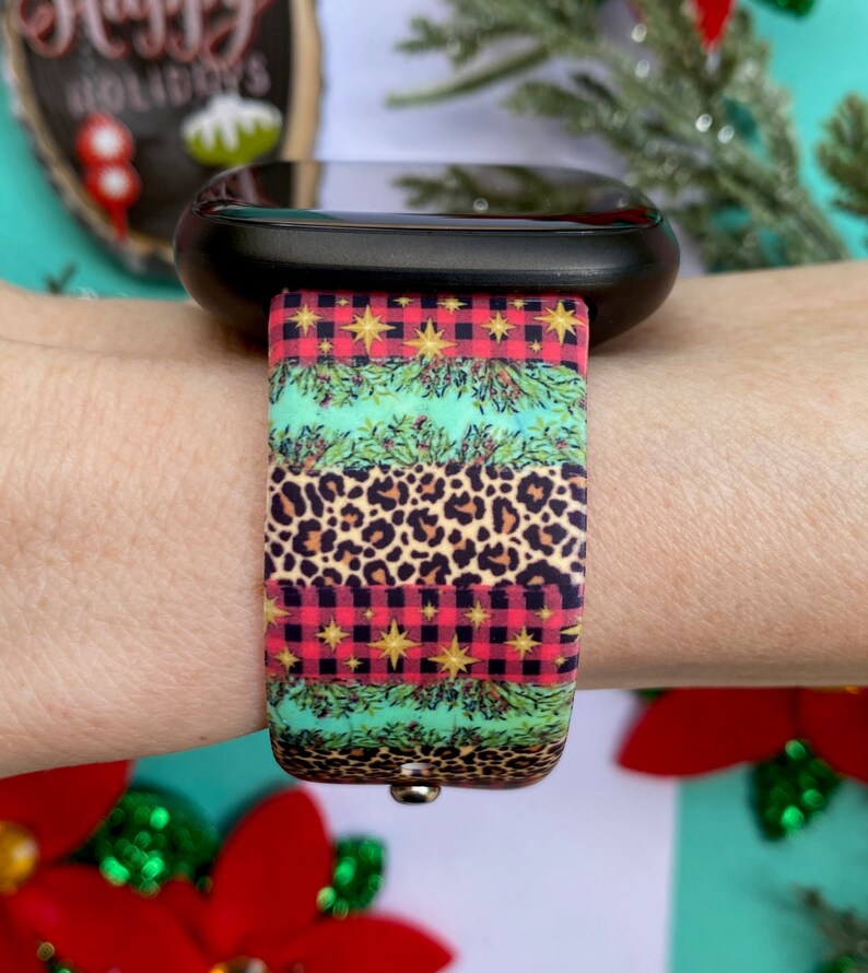 Op de afbeelding: Een zwarte smartwatch met een kleurrijk kerstbandje. Het bandje heeft een rood-zwart ruitpatroon, een groen-gouden bladpatroon en een luipaardprint.