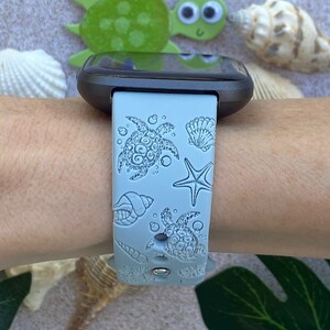 May include: A light blue silicone watch band with a sea turtle and seashell design. The band is on a person's wrist.