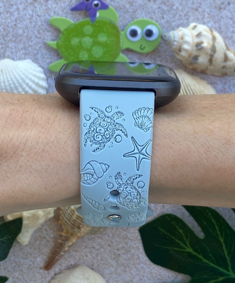 May include: A light blue silicone watch band with a sea turtle and seashell design. The band is on a person's wrist.