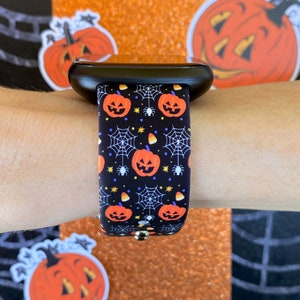 K&ouml;nnte beinhalten: Eine schwarze Smartwatch mit einem Halloween-Armband. Das Armband zeigt ein sich wiederholendes Muster aus orangefarbenen K&uuml;rbissen, Spinnweben und Maisbonbons auf schwarzem Hintergrund.