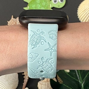 May include: A light blue watch band with a sea turtle and seashell design. The band is attached to a black watch face. The design includes two sea turtles, a starfish, and various seashells. The band is made of a soft, flexible material.