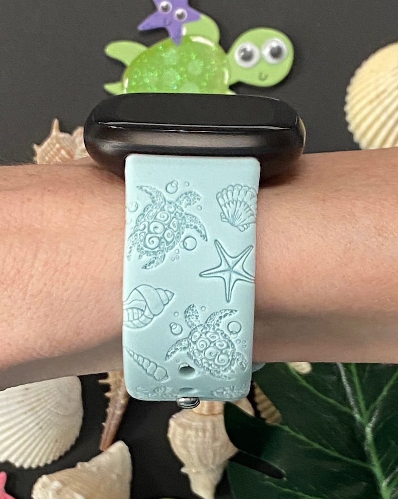 May include: A light blue watch band with a sea turtle and seashell design. The band is attached to a black watch face. The design includes two sea turtles, a starfish, and various seashells. The band is made of a soft, flexible material.