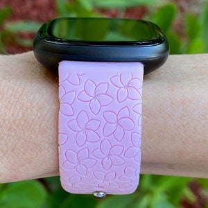 May include: A pink silicone watch band with a floral pattern. The band is designed for a smartwatch with a black face.