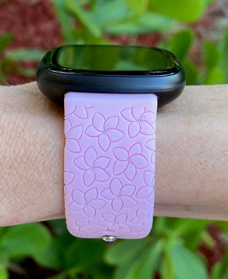 May include: A pink silicone watch band with a floral pattern. The band is designed for a smartwatch with a black face.