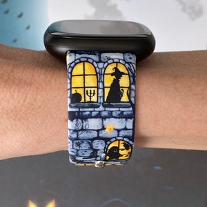 May include: A black smartwatch with a blue and yellow Halloween-themed band. The band features a brick wall with windows and silhouettes of a witch, a cat, and a ghost.