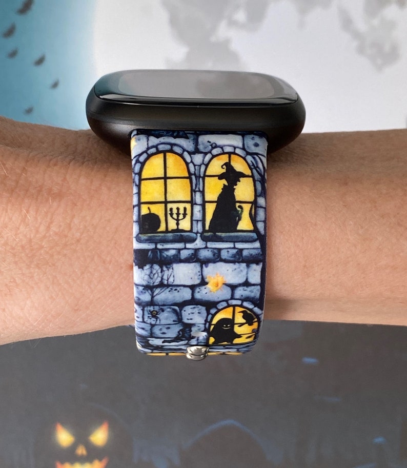 May include: A black smartwatch with a blue and yellow Halloween-themed band. The band features a brick wall with windows and silhouettes of a witch, a cat, and a ghost.