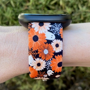 May include: A black smartwatch with a bright orange, white and black patterned band featuring flowers, ghosts and spiderwebs.
