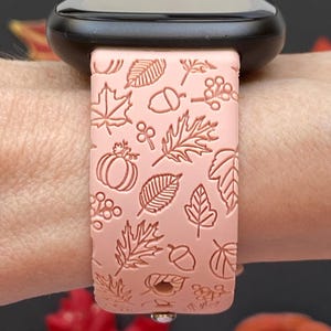May include: A light pink watch band with a fall-themed design. The band features embossed images of leaves, acorns, berries, and a pumpkin. The watch band is attached to a black smartwatch.