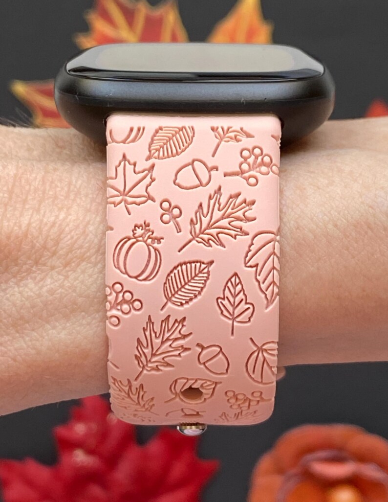 May include: A light pink watch band with a fall-themed design. The band features embossed images of leaves, acorns, berries, and a pumpkin. The watch band is attached to a black smartwatch.