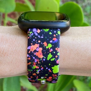 Puede incluir: Un reloj inteligente negro con una banda de dise&ntilde;o de pintura salpicada de colores. La banda tiene salpicaduras de pintura verde, naranja, rosa y p&uacute;rpura sobre un fondo negro.
