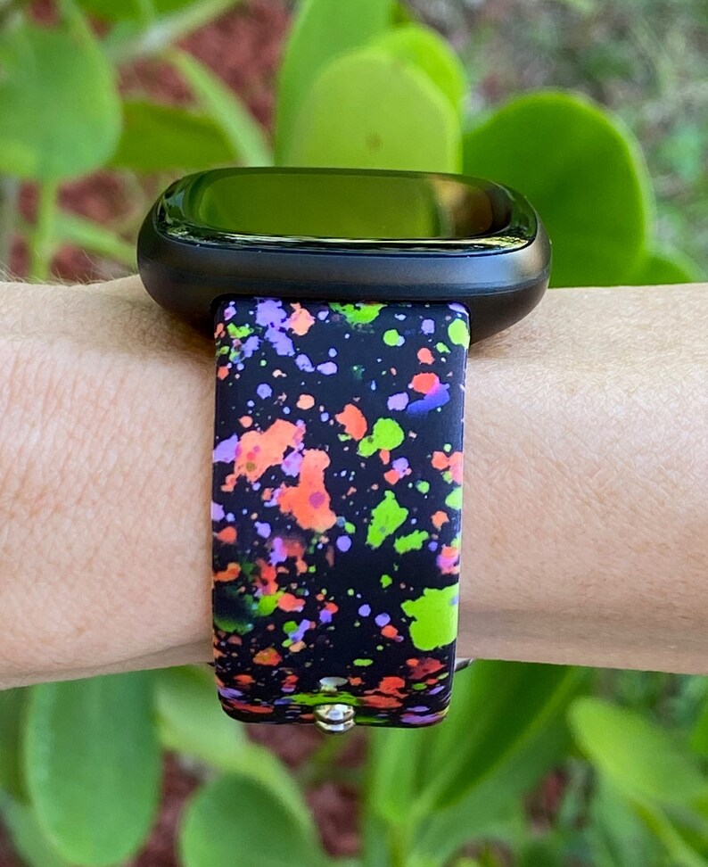 Puede incluir: Un reloj inteligente negro con una banda de dise&ntilde;o de pintura salpicada de colores. La banda tiene salpicaduras de pintura verde, naranja, rosa y p&uacute;rpura sobre un fondo negro.