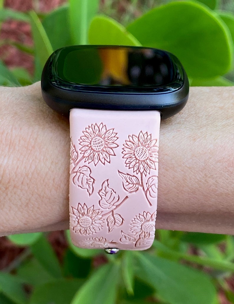 May include: A pink silicone watch band with a floral design featuring white sunflowers and leaves. The band is designed for a smartwatch.