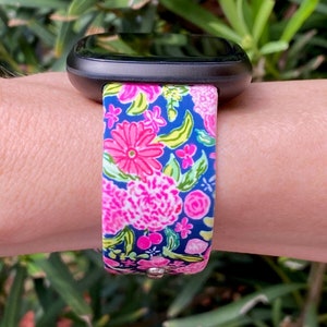 Pu&ograve; includere: Uno smartwatch nero con un cinturino a fantasia floreale. Il cinturino &egrave; blu con fiori rosa, bianchi e verdi.