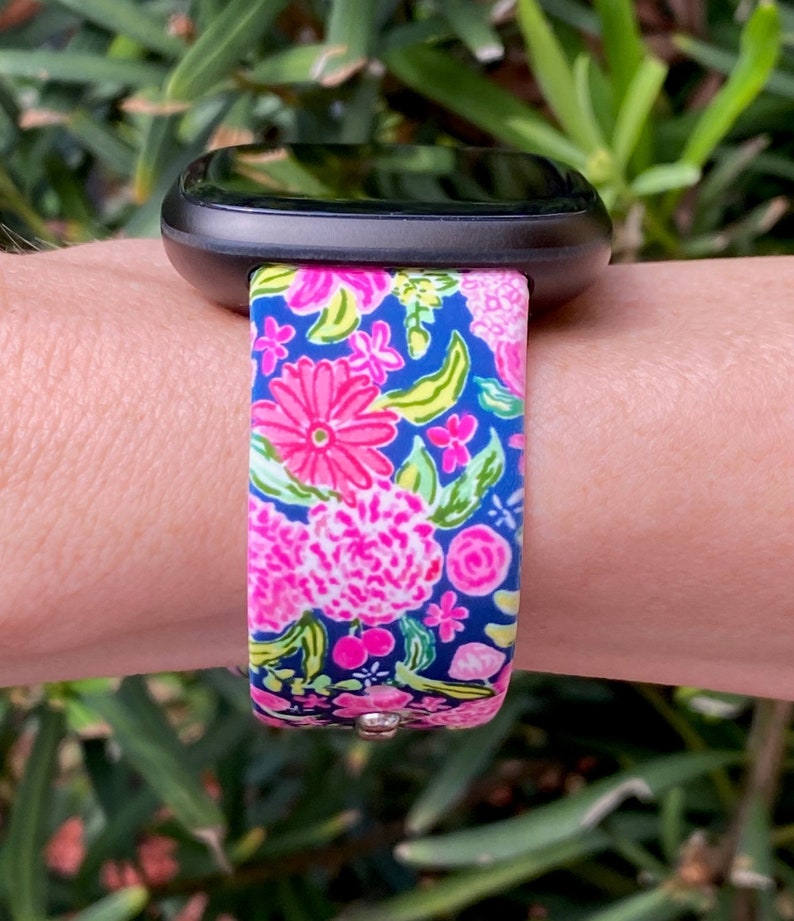 Pu&ograve; includere: Uno smartwatch nero con un cinturino a fantasia floreale. Il cinturino &egrave; blu con fiori rosa, bianchi e verdi.