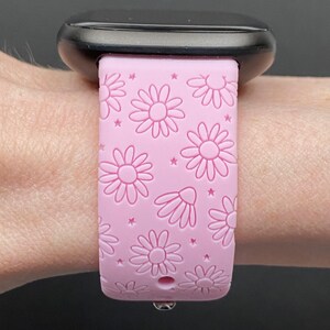May include: A pink silicone watch band with a daisy print. The band is designed for a smartwatch and has a small hole for the watch's charging port.