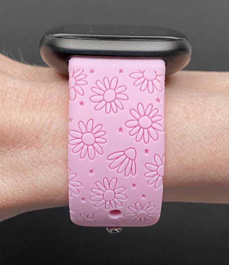 May include: A pink silicone watch band with a daisy print. The band is designed for a smartwatch and has a small hole for the watch's charging port.