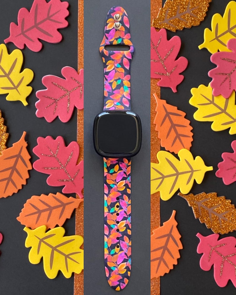 Pu&ograve; includere: Uno smartwatch nero con un cinturino a fantasia di foglie colorate. Il cinturino presenta un design di foglie arancioni, rosa e gialle su sfondo blu scuro. Ritagli decorativi di foglie a tema autunnale circondano l'orologio.