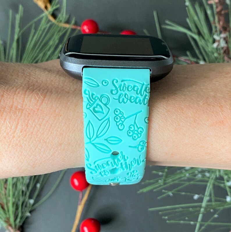 Pu&ograve; includere: Un cinturino per smartwatch color turchese con un designo bianco che presenta foglie, bacche e il testo "Sweater Weather".