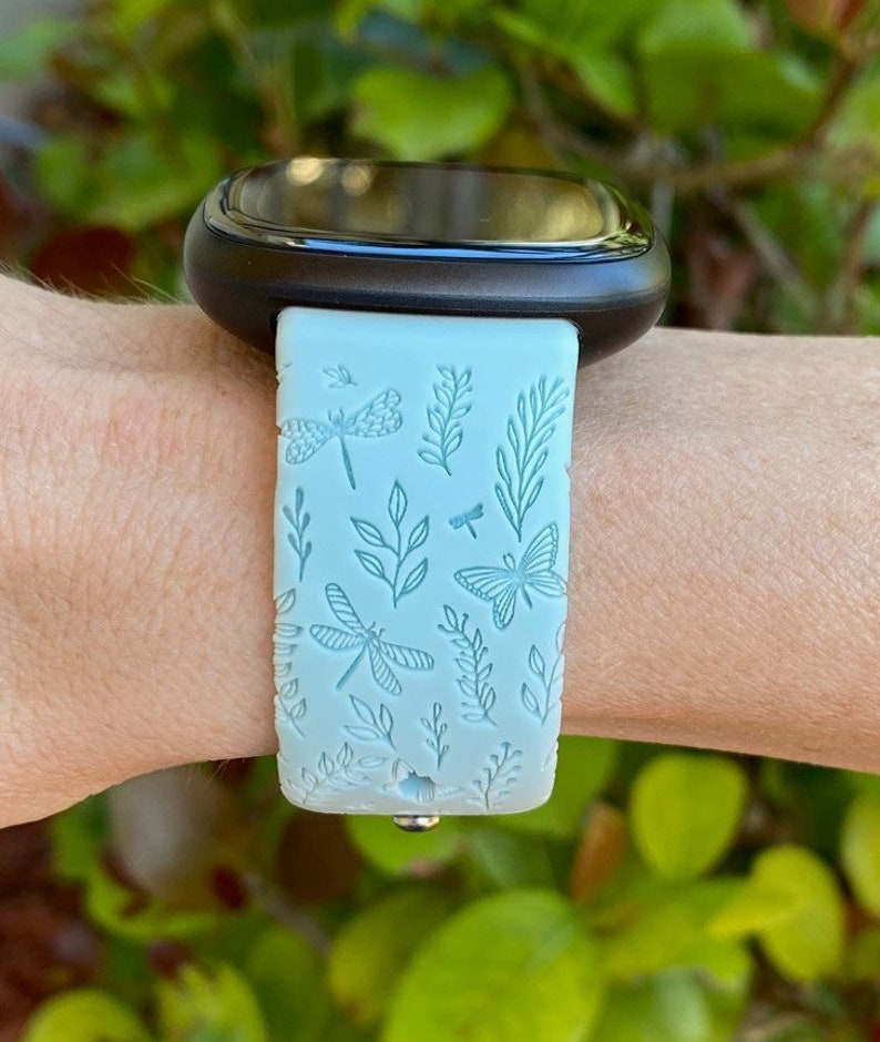 May include: A light blue watch band with a dragonfly and butterfly design. The band is made of silicone and has a smooth finish.