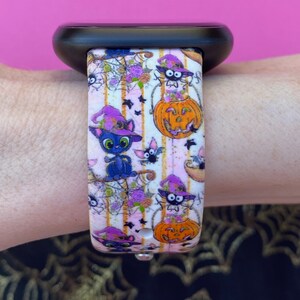 以下が含まれることがあります： ハロウィンテーマのピンクと白のパターンのApple Watchバンド。バンドには、魔女の帽子をかぶった黒い猫、コウモリ、カボチャ、クモの巣が描かれています。