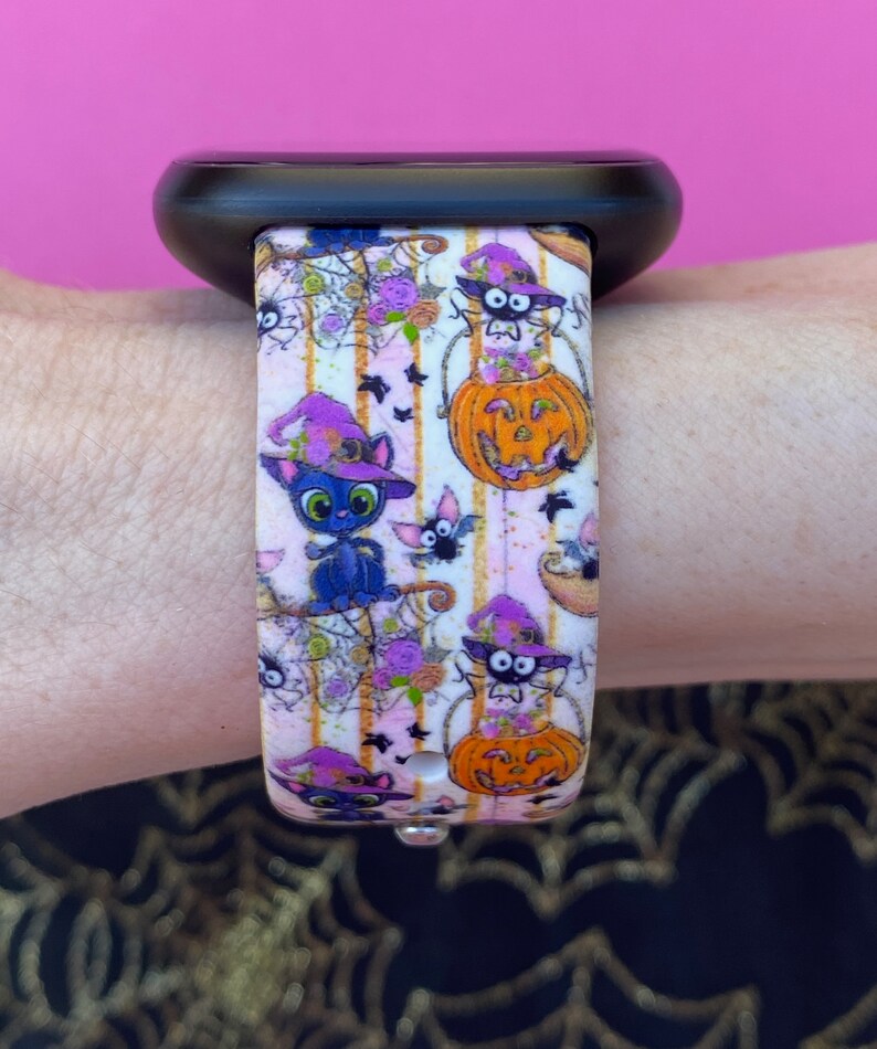 以下が含まれることがあります： ハロウィンテーマのピンクと白のパターンのApple Watchバンド。バンドには、魔女の帽子をかぶった黒い猫、コウモリ、カボチャ、クモの巣が描かれています。