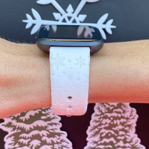 K&ouml;nnte beinhalten: Wei&szlig;e Smartwatch mit einem Armband mit Schneeflockenmuster. Das Zifferblatt der Uhr ist schwarz und das Armband ist wei&szlig; mit einem Schneeflockenmuster.