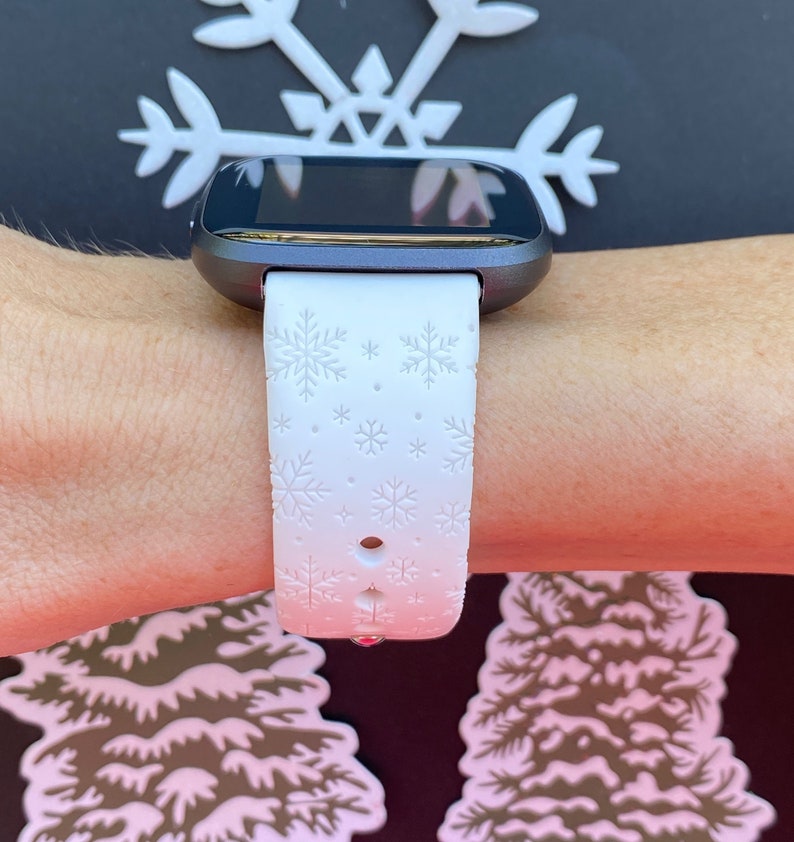 K&ouml;nnte beinhalten: Wei&szlig;e Smartwatch mit einem Armband mit Schneeflockenmuster. Das Zifferblatt der Uhr ist schwarz und das Armband ist wei&szlig; mit einem Schneeflockenmuster.