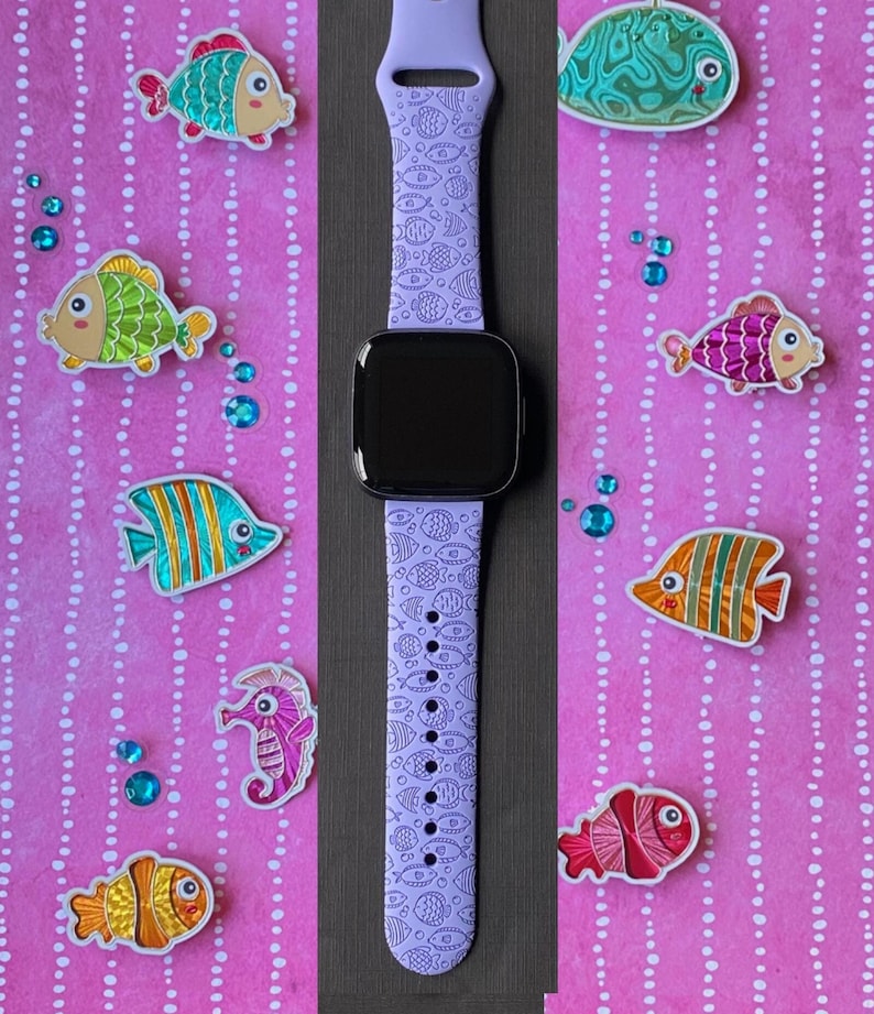 May include: A purple watch strap with a white and blue fish pattern. The strap is on a pink background with colourful fish stickers.