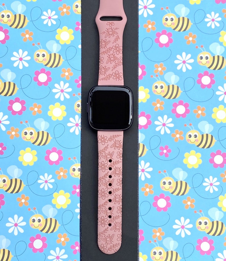 K&ouml;nnte beinhalten: Ein staubrosa Smartwatch-Armband mit einem Blumen- und Bienenmuster. Das Armband ist mit einem schwarzen Smartwatch-Zifferblatt kombiniert. Der Hintergrund ist ein hellblaues Muster mit Bienen und Blumen.
