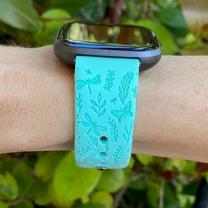 K&ouml;nnte beinhalten: Ein t&uuml;rkisfarbenes Armband f&uuml;r eine Smartwatch mit einem Libellen- und Schmetterlingsmuster. Das Armband ist aus Silikon und hat eine strukturierte Oberfl&auml;che.