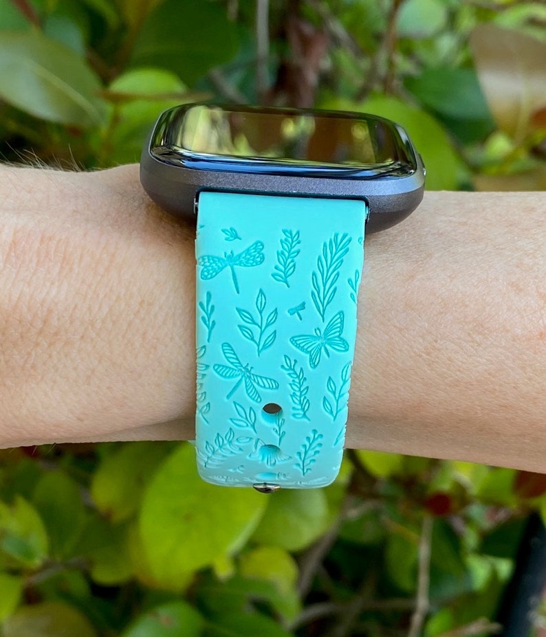 K&ouml;nnte beinhalten: Ein t&uuml;rkisfarbenes Armband f&uuml;r eine Smartwatch mit einem Libellen- und Schmetterlingsmuster. Das Armband ist aus Silikon und hat eine strukturierte Oberfl&auml;che.