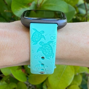 Op de afbeelding: Een turquoise smartwatch band met een gedetailleerd zeeschildpad ontwerp. De band is gemaakt van siliconen en heeft een zwart horloge gezicht.