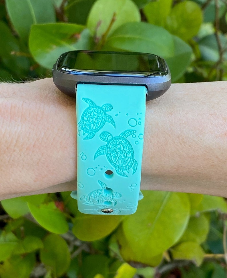 Op de afbeelding: Een turquoise smartwatch band met een gedetailleerd zeeschildpad ontwerp. De band is gemaakt van siliconen en heeft een zwart horloge gezicht.
