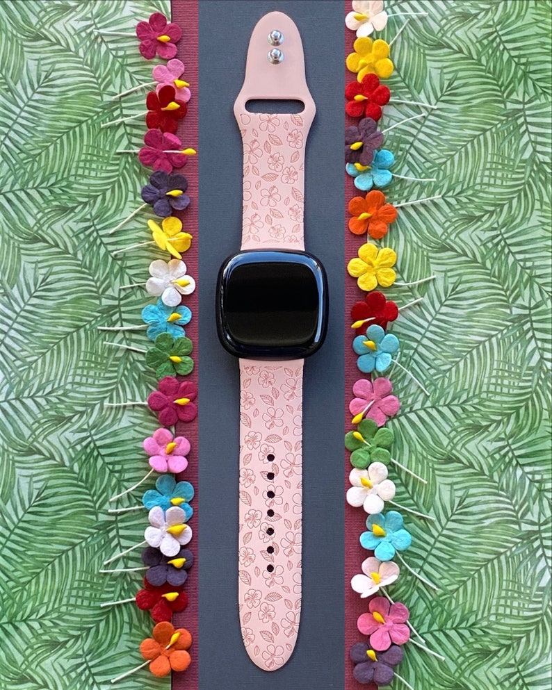 May include: A pink Apple Watch band with a floral pattern. The band is on a black background with colourful paper flowers on either side.