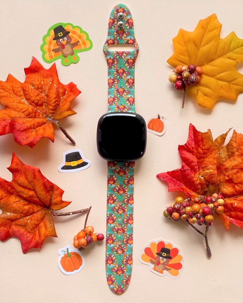 K&ouml;nnte beinhalten: Ein t&uuml;rkisfarbenes und orangefarbenes Armband mit einem schwarzen Zifferblatt. Das Armband zeigt ein sich wiederholendes Muster aus Truth&auml;hnen mit Pilgerh&uuml;ten. Die Uhr ist von Herbstdekorationen umgeben, darunter Bl&auml;tter, K&uuml;rbisse und Beeren.